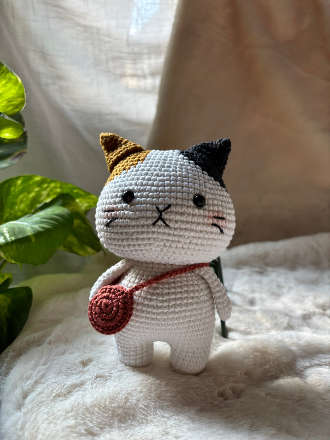 Crochet calico cat with red crossbody bag beside a plant