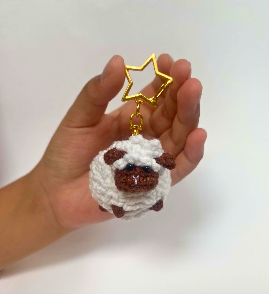 White crochet sheep keychain with brown face on a gold star clasp, held in a hand