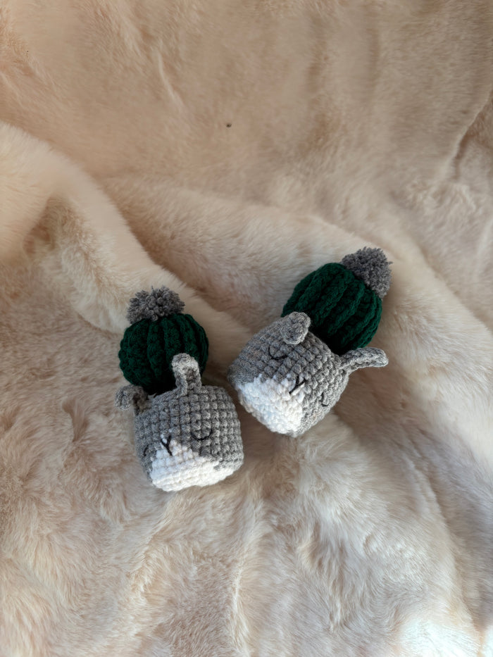 Two crochet cacti with grey pom-poms in grey cat pots lying down