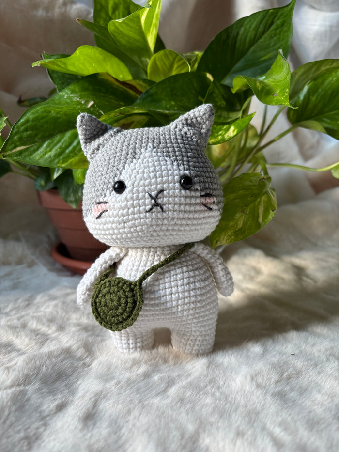 Grey and white crochet cat with green crossbody bag beside a plant