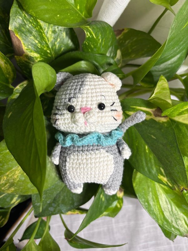 Crocheted grey and white kitten with a light-blue ruffled collar, waving among green leaves